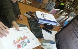 Chłopiec podsuwa książkę pod czytnik przy biurku aby zeskanować kod. W tle widać dzieci w kolejce.