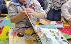 Chłopiec siedzi na kocyku i przegląda książeczkę. W tle widać inne dzieci