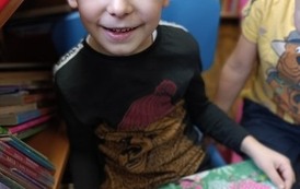 Chłopiec o blond włosach trzyma w ręku książkę i uśmiecha się.