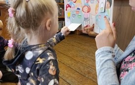 Bibliotekarka trzyma otwartą książkę dla dzieci, dziewczynka stoi przed książką i otwiera okienko w książce.