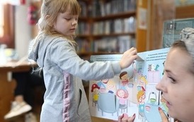 Bibliotekarka trzyma otwartą książkę dla dzieci, dziewczynka stoi przed książką i otwiera okienko w książce.
