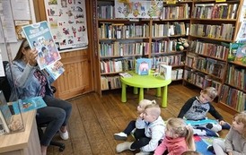 Dzieci siedzą na kocykach, Pani bibliotekarka pokazuje im tytułową okładkę książki.