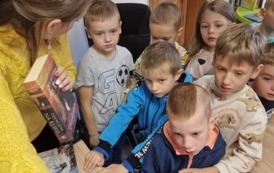 Dzieci stoją przy biurku, a Pani bibliotekarka trzyma w ręku książkę i im pokazuje coś na komputerze.