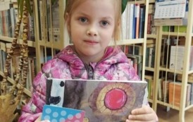 Dziewczynka pokazuje swoją nową książeczkę i kartę małego czytelnika. W tle jest kwiat doniczkowy i p&oacute;łki z książkami w bibliotece w Zblewie.