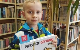 Chłopiec pokazuje swoją nową książeczkę,  w tle widać kwiat doniczkowy i p&oacute;łki z książkami w bibliotece w Zblewie.