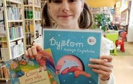 Dziewczynka stoi i pokzuje sw&oacute;j dyplom Małego Czytelnika i w prawej ręce trzyma jeszcze wypożyczoną książeczkę