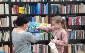 Dziewczynka stoi przy bibliotekarce i rączką wyjmuje rybkę z pluszowej książeczki, ktorą trzyma bibliotekarka. 