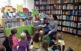 Dzieci siedzą na dywanie na przeciwko bibliotekarki i patrzą jak bibliotekarka pokazuje im książeczkę pluszową.