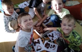 Siedmiu chłopc&oacute;w siedzi na dywanie i układają z kasztan&oacute;w wzory na kartce.Ich twarze uśmiechnięte są skierowane do aparatu fotograficznego. 