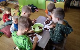 Czterech chłopc&oacute;w siedzi przy stolku i układa wzory z kasztan&oacute;w, w tle widać innych chłopc&oacute;w siedzących na dywanie.