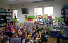 Dzieci siedzą na dywanie i mają ręce w g&oacute;rze naśladując to co pokazuje Pani bibliotekarka, kt&oacute;ra siedzi na przeciwko nich.