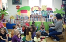 Dzieci siedzą na dywanie i patrzą na Panią bibliotekarkę, kt&oacute;ra czyta im bajkę.
