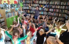 Dzieci siedzą na dywanie i mają ręce w g&oacute;rze naśladując to co pokazuje Pani bibliotekarka, kt&oacute;ra siedzi na przeciwko nich.