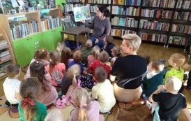Dzieci i opiekunka siedzą na dywanie i słuchają bajki czytanej przez bibliotekarkę.