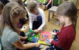 Trzy dziewczynki siedzą w bibliotece na podłodze i przeglądają pluszową książeczkę.