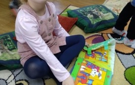 Dziewczynka o blond włosach siedzi w bibliotece na dywanie i ogląda pluszową książeczkę.