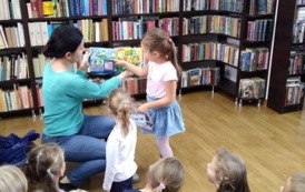 Dziewczynka stoi przy bibliotekarce i rączką wyjmuje rybkę z pluszowej książeczki, ktorą trzyma bibliotekarka. Reszta dzieci siedzi na dywanie i patrzy.