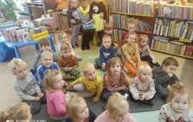 Grupa dzieci siedzi na dywanie skierowana twarzą do aparatu, a po środku siedzi bibliotekarka i obejmuje dwie dziewczynki.