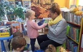 Pani bibliotekarka trzyma pluszową książeczkę i stoi przy niej dzieczynka w r&oacute;żowym sweterku i pokazuje coś rączką w książeczce.  Widać kilkoro dzieci.