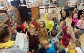 Dzieci siedzą na dywanie, niekt&oacute;re z nich zgłaszają się aby odpowiedzieć na zagadkę powiedzianą przez Panią bilbiotekarkę.Bibliotekarka siedzi na krzesełku po środku i czyta z kartki zagadki.