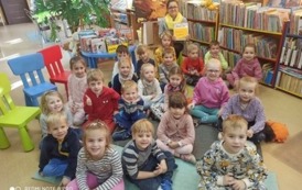 Grupa dzieci siedząca na dywanie, z tyłu Pani bibliotekarka z książką w rękach - pozują do zdjęcia grupowego.