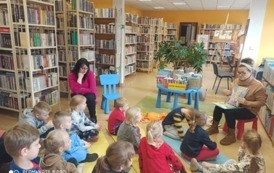 Dzieci siedzą na karimatach i słuchają bajkę czytaną przez bibliotekarkę. 