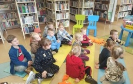 Dzieci siedzą na karimatach i słuchają bajkę czytaną przez bibliotekarkę.