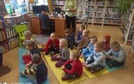 Dzieci siedzą na karimatach i słuchają czytanej przez bibliotekarkę bajki. W tle widać Panią opiekunkę klasy.