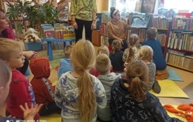 Dzieci siedzą na dywanie i słuchają bajkę czytaną przez bibliotekarkę. W tle widać stojącą Panią opiekunkę klasy.