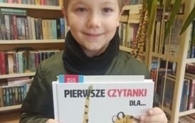 Chłopiec o blond włosach stoi ze swoją wyprawką czytelniczą w bibliotece w Zblewie
