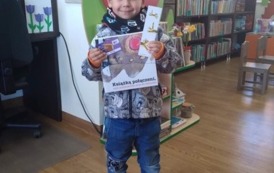 Chłopiec o blond włosach stoi w bibliotece w Pinczynie z nową książką, zakładką do ksiązki i kartą Małego Czytelnika
