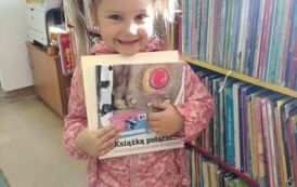 Dziewczynka w r&oacute;żowej kurtce i dw&oacute;ch kitkach stoi przy regale z książkami trzymając swoją nową książkę i kartę Małego Czytelnika