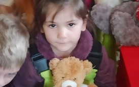 Dziewczynka w ciemnych włosach trzyma maskotkę  brązowego misia. W tle widać inne dzieci.