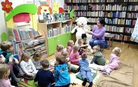 Dzieci siedzą na dywanie i poduszkach i słuchają bajki czytanej przez bibliotekarkę. Z lewej strony siedzi na krzesle duży biały miś.