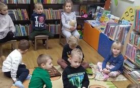 Dzieci siedża na dywanie i krzesełkach i słuchają bajki czytanej przez bibliotekarkę.