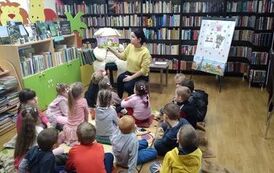 Dzieci siedzą w grupie na dywanie i słuchają bajki, kt&oacute;rą czyta bibliotekarka.Obok widać siedzącego misia,a  z prawej strony stoi przystrojona na Dzień Misia magnetyczna tablica.