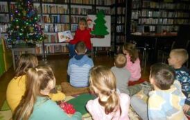 Dzieci siedzą na poduszkach i słuchają bibliotekarki, kt&oacute;ra czyta im bajkę.