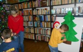 Chłopiec stoi przy tablicy na kt&oacute;rej przyczepiona jest choinka filcowa i coś do niej przylepia. Obok stoi bibliotekarka i coś szuka w woreczku i obok stoi jeszcze chłopiec.