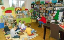 Dzieci siedzą na dywanie,a bibliotekarka czyta im bajkę. Z prawej strony stoi przystrojona choinka.
