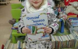 Dziewczynka w r&oacute;żowej czapce pokazuje sw&oacute;j dyplom Małego Czytelnika, w rączce też trzyma kredki i kartę magnetyczną. 