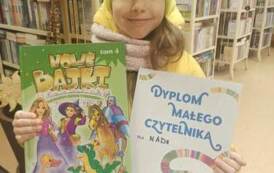 Dziewczynka w ż&oacute;łtej czapce pokazuje swoją wypożyczoną książeczkę i dyplom Małego Czytelnika