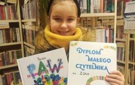 Dziewczynka o blond włosach pokazuje sw&oacute;j Dyplom Małego Czytelnika i wypożyczoną książeczkę