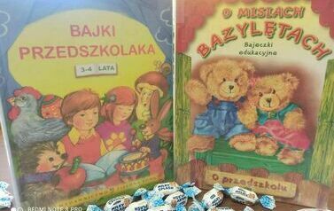 Na stole są postawione dwie książki dla dzieci: Bajki Przedszkolaka i O Misiach Bazylentach. Przy książkach leżą cukierki.