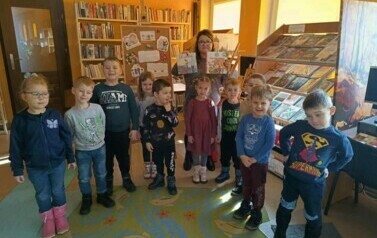 Dzieci stoją w grupie na dywanie, za nimi stoi bibliotekarka trzymając w ręku książeczki o babci i dziadku.