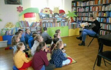 Dzieci siedzą na dywanie i słuchają bajki, kt&oacute;rą czyta im bibliotekarka.