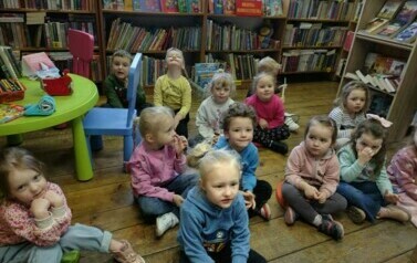 Dzieci siedzą na siedziskach i słuchają bajki, kt&oacute;rą czyta im bibliotekarka.