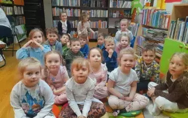 Dzieci siedzą na dywanie i siedziskach i słuchają bajki, kt&oacute;rą czyta im bibliotekarka.
