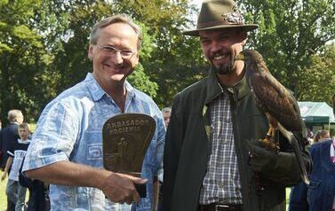 Wojciech Cejrowski stoi z Leśnikiem, kt&oacute;ry trzyma w ręku sokoła. Pan Cejrowski trzyma statuetkę Ambasador Kociewia