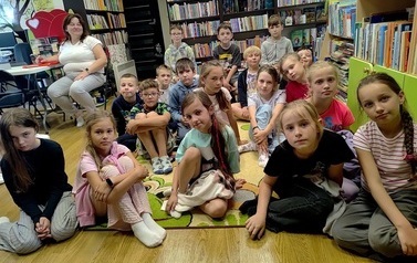 Dzieci siedzą na dywanie i słuchają czytanki, po lewej stronie na krześle siedzi nauczycielka.