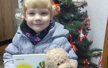 Uśmiechnięta dziewczynka w blond włosach trzyma misia, nową książkę i kartę Małego Czytelnika. Za nią stoi ustrojona choinka.
